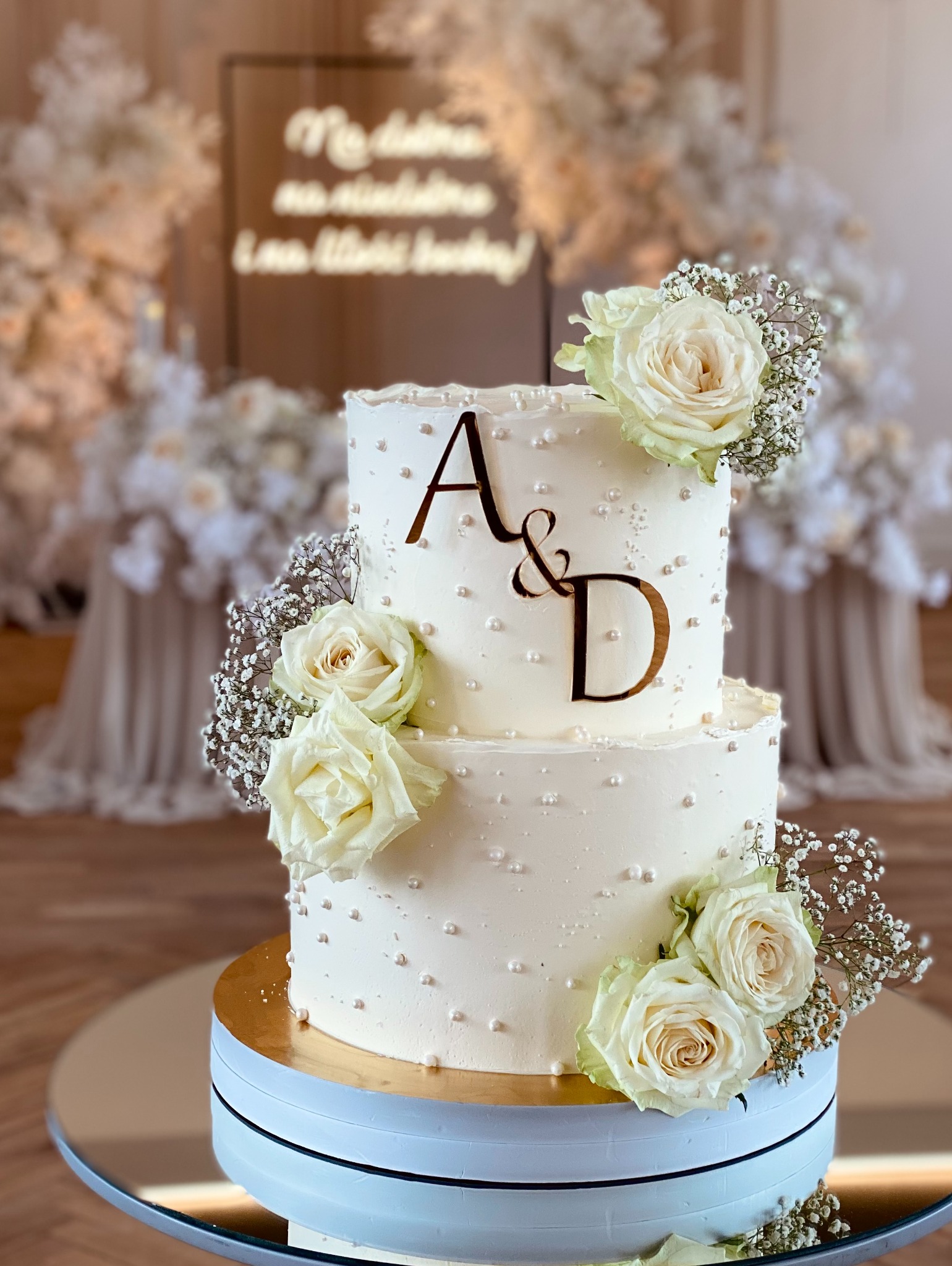Dwupiętrowy tort weselny w kremowej tonacji, zdobiony białymi różami i perłowymi koralikami, z inicjałami 'A&D' na tle dekoracji kwiatowych i napisu.