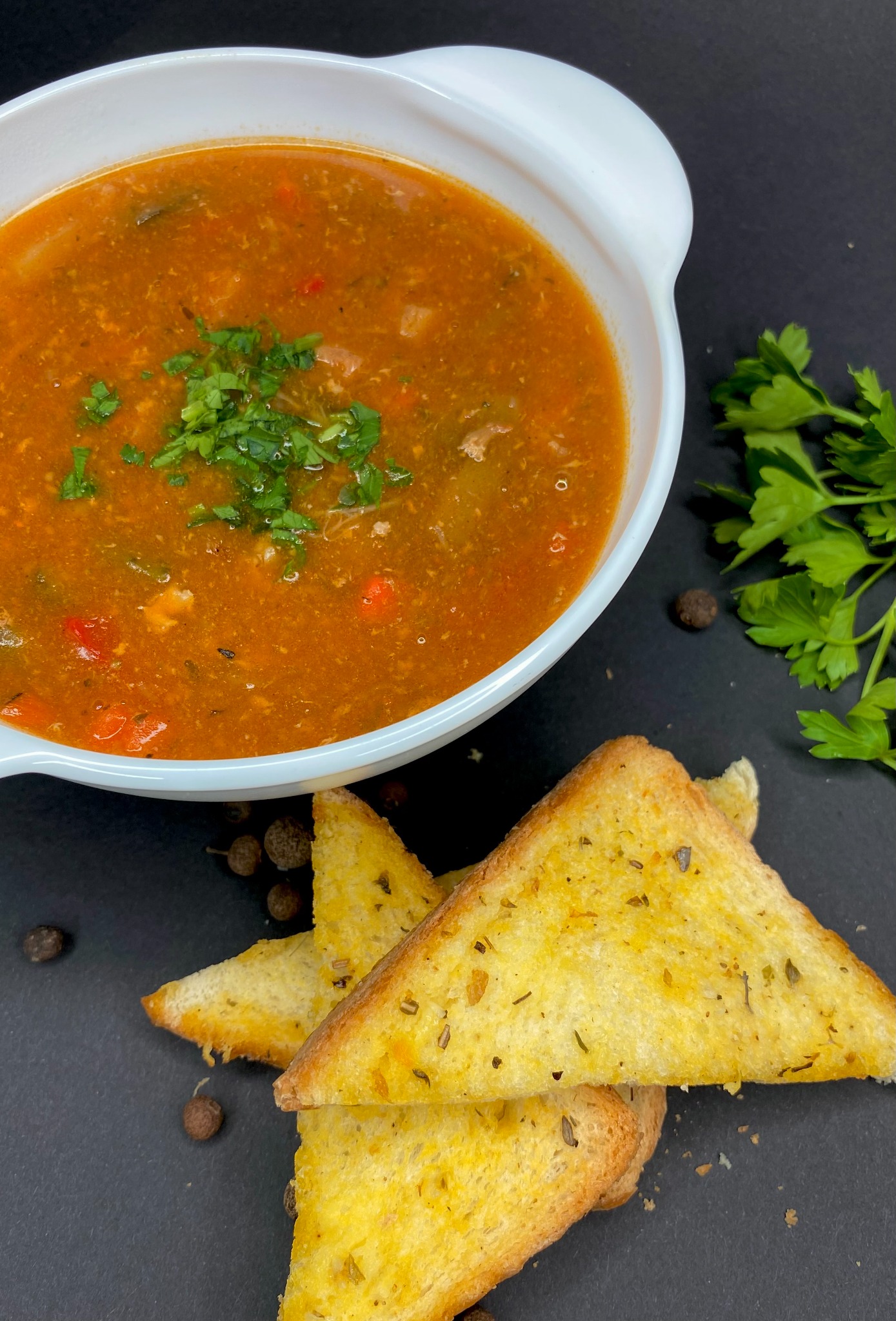 Apetyczna zupa warzywna w białej misce, posypana świeżą pietruszką, podana z grzankami czosnkowymi na ciemnym tle. Idealna propozycja na catering.