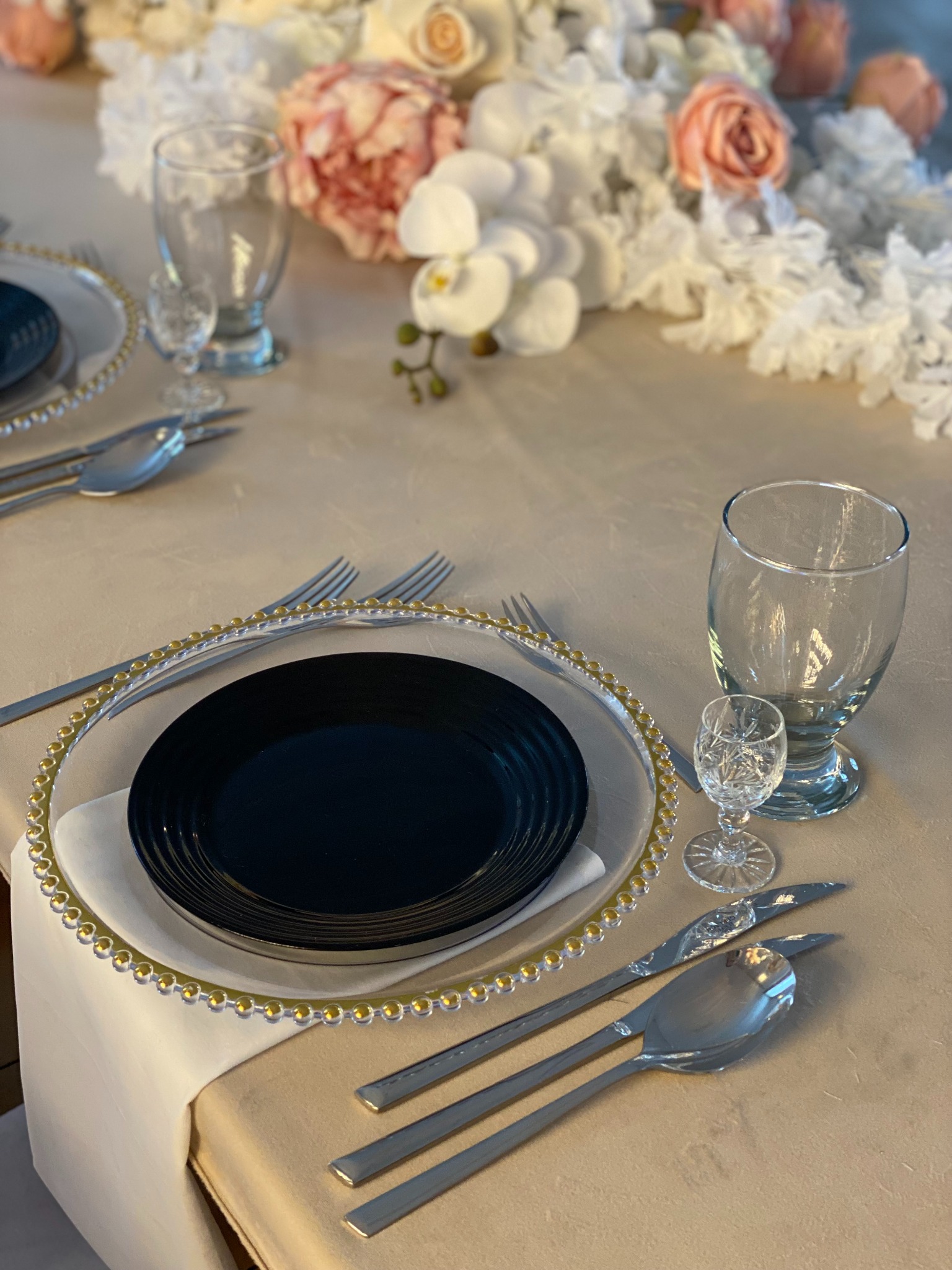 Elegancko nakryty stół z czarnym talerzem, srebrnymi sztućcami i kryształowymi kieliszkami, udekorowany kwiatami. Detal aranżacji stołu na przyjęcie.