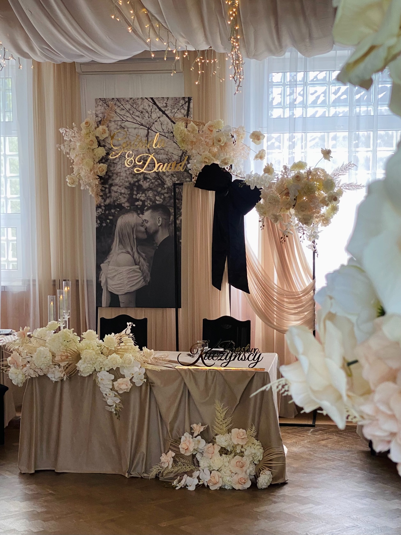 Elegancko udekorowany stół weselny z kwiatami w odcieniach bieli i beżu, za nim czarno-białe zdjęcie pary młodej i złoty napis. Całość w jasnej, przestronnej sali.
