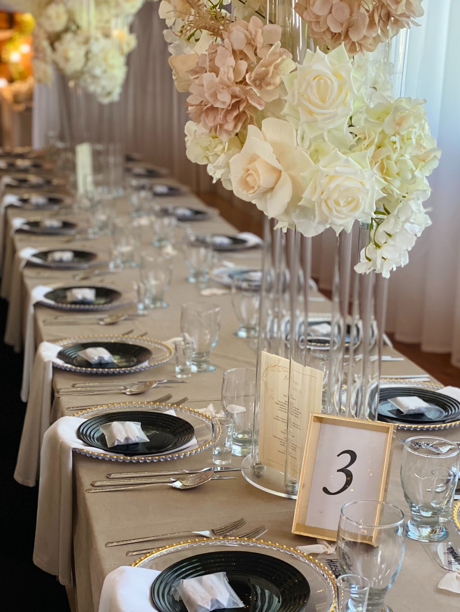 Elegancko nakryty stół z kwiatowymi dekoracjami, czarnymi talerzami ze złotymi obwódkami, szkłem i numerem stołu '3'. Idealne na przyjęcie weselne lub firmowe.