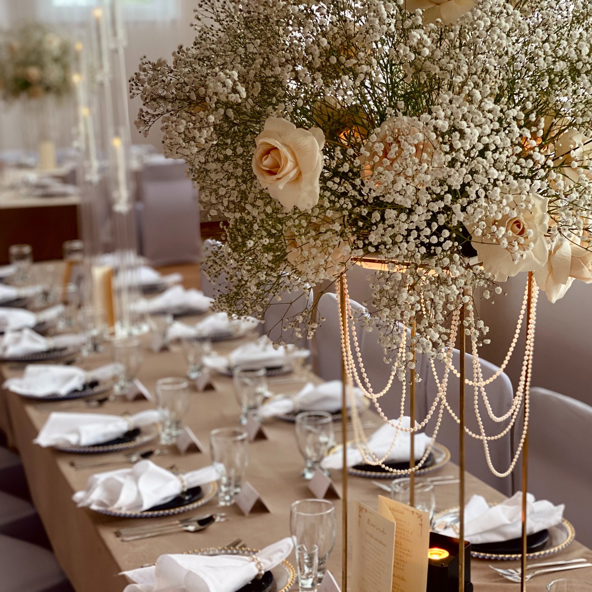 Elegancka dekoracja stołu z wysokim bukietem z białych róż i gipsówki, perłowe girlandy, czarne talerze ze złotym obramowaniem, nakrycia. Subtelne oświetlenie, uroczysty nastrój.