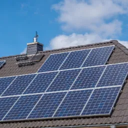 Panele słoneczne na brązowym dachu domu jednorodzinnego, widoczne komin z metalową ozdobą i błękitne niebo z chmurami w tle.