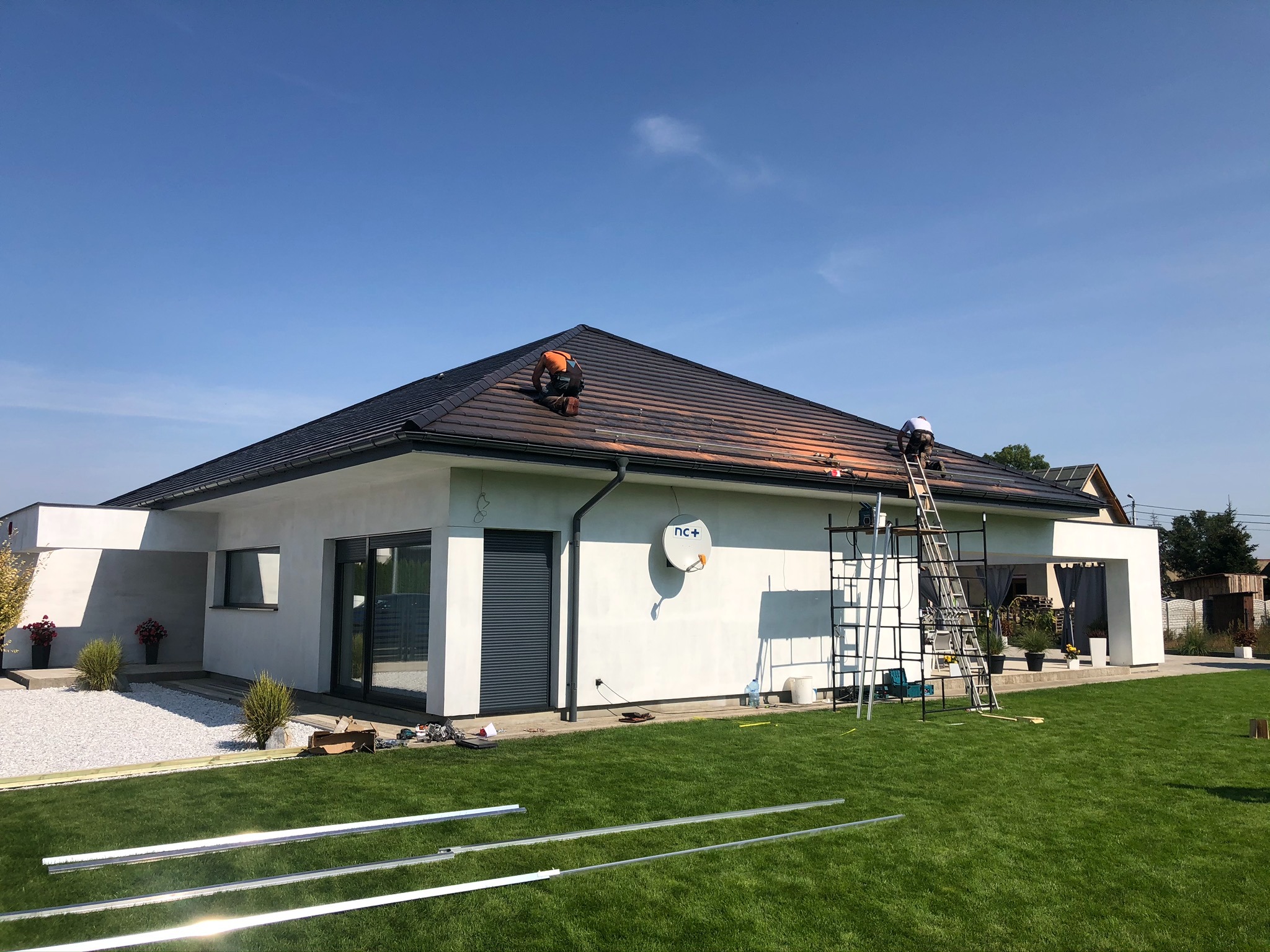 Montaż paneli na dachu nowoczesnego domu jednorodzinnego z widoczną anteną satelitarną, aluminiowe profile leżą na trawniku.