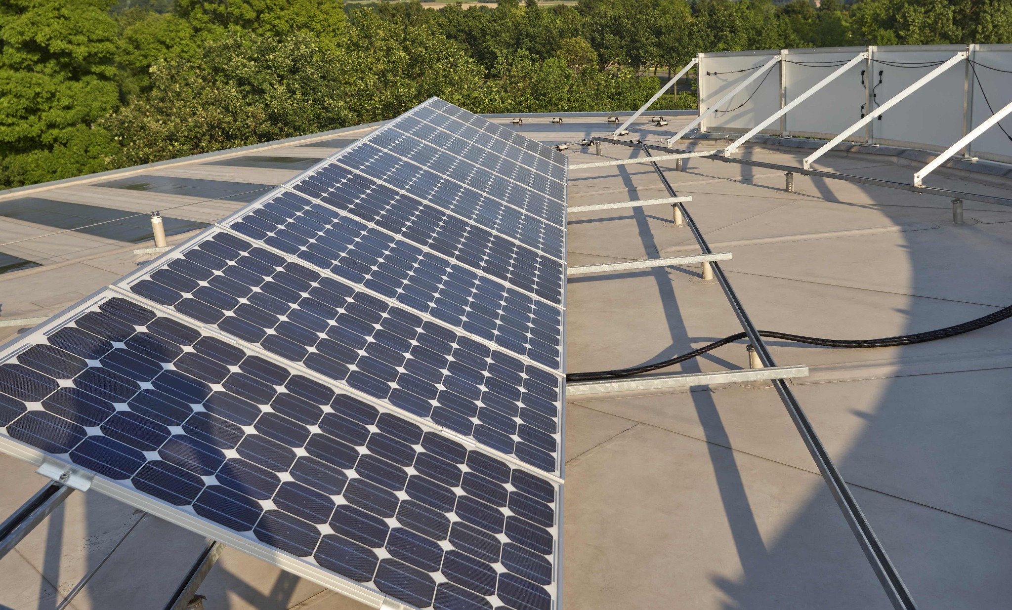 Instalacja paneli słonecznych na płaskim dachu z widocznym systemem montażowym i zielenią w tle, ujęcie z góry.