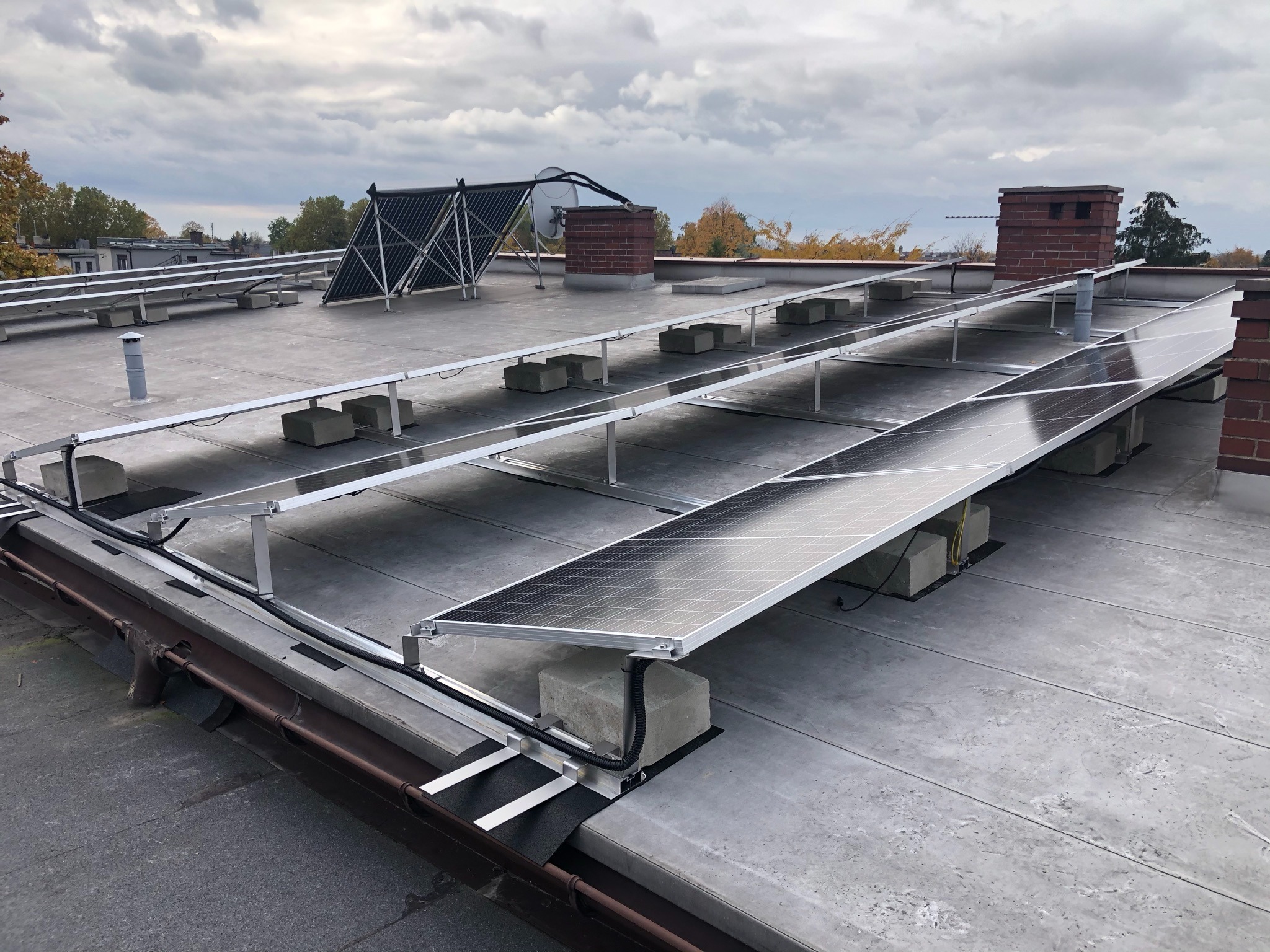 Instalacja fotowoltaiczna i kolektory słoneczne na płaskim dachu z szarym pokryciem, panele zamontowane na betonowych blokach, w tle ceglany komin i pochmurne niebo.