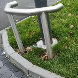 Srebrna, metalowa barierka ochronna przy chodniku w Częstochowie, otoczona kostką brukową i trawnikiem z opadłymi liśćmi.