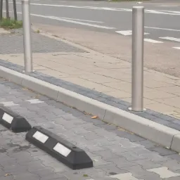 Słupki ze stali nierdzewnej oddzielające chodnik od jezdni, z odbojnikami parkingowymi na pierwszym planie, ułożone na szarej kostce brukowej.