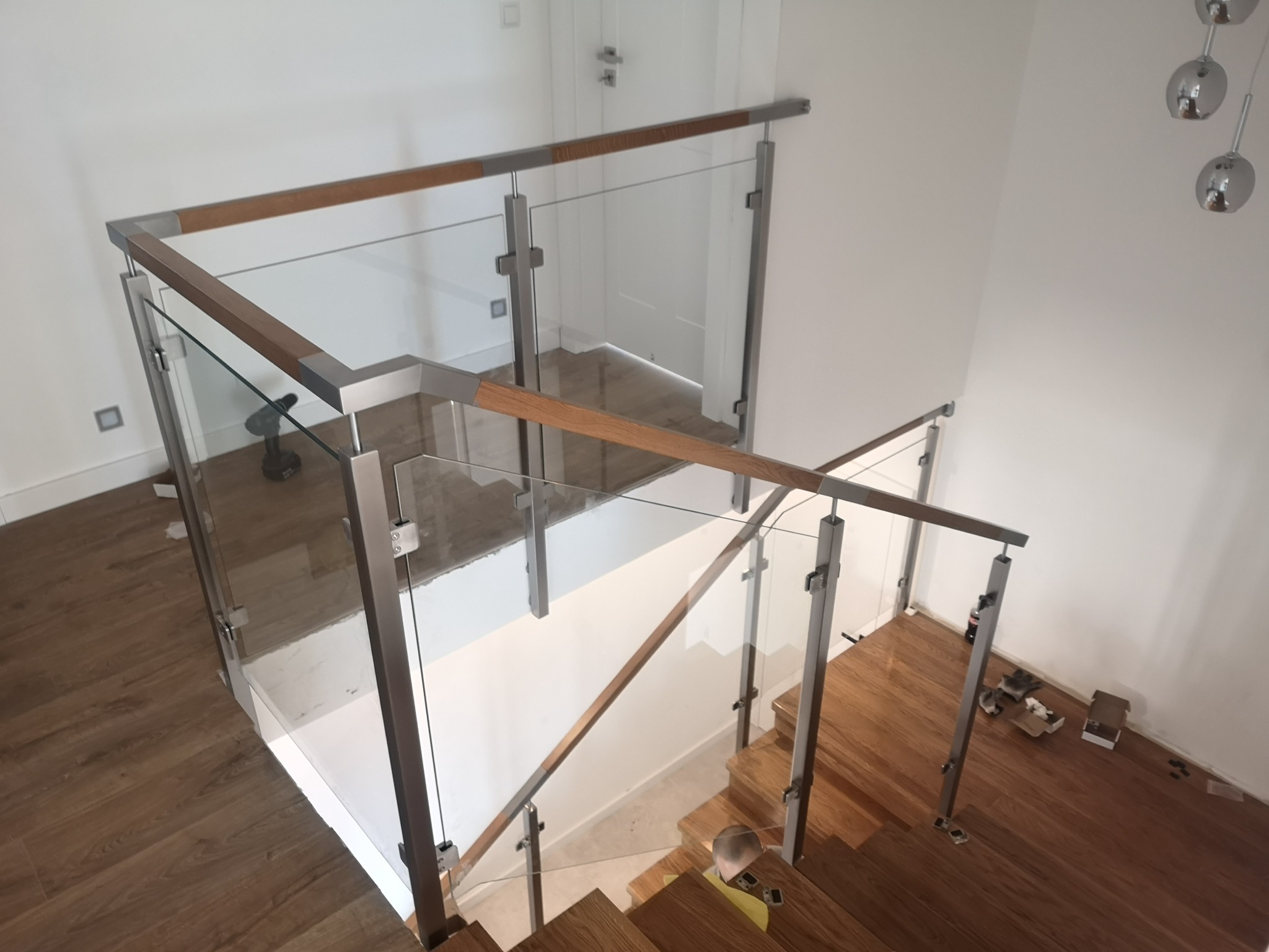 Widok z góry na nowoczesne schody z drewnianymi stopniami i szklaną balustradą z drewnianą poręczą, montowaną do metalowych słupków.