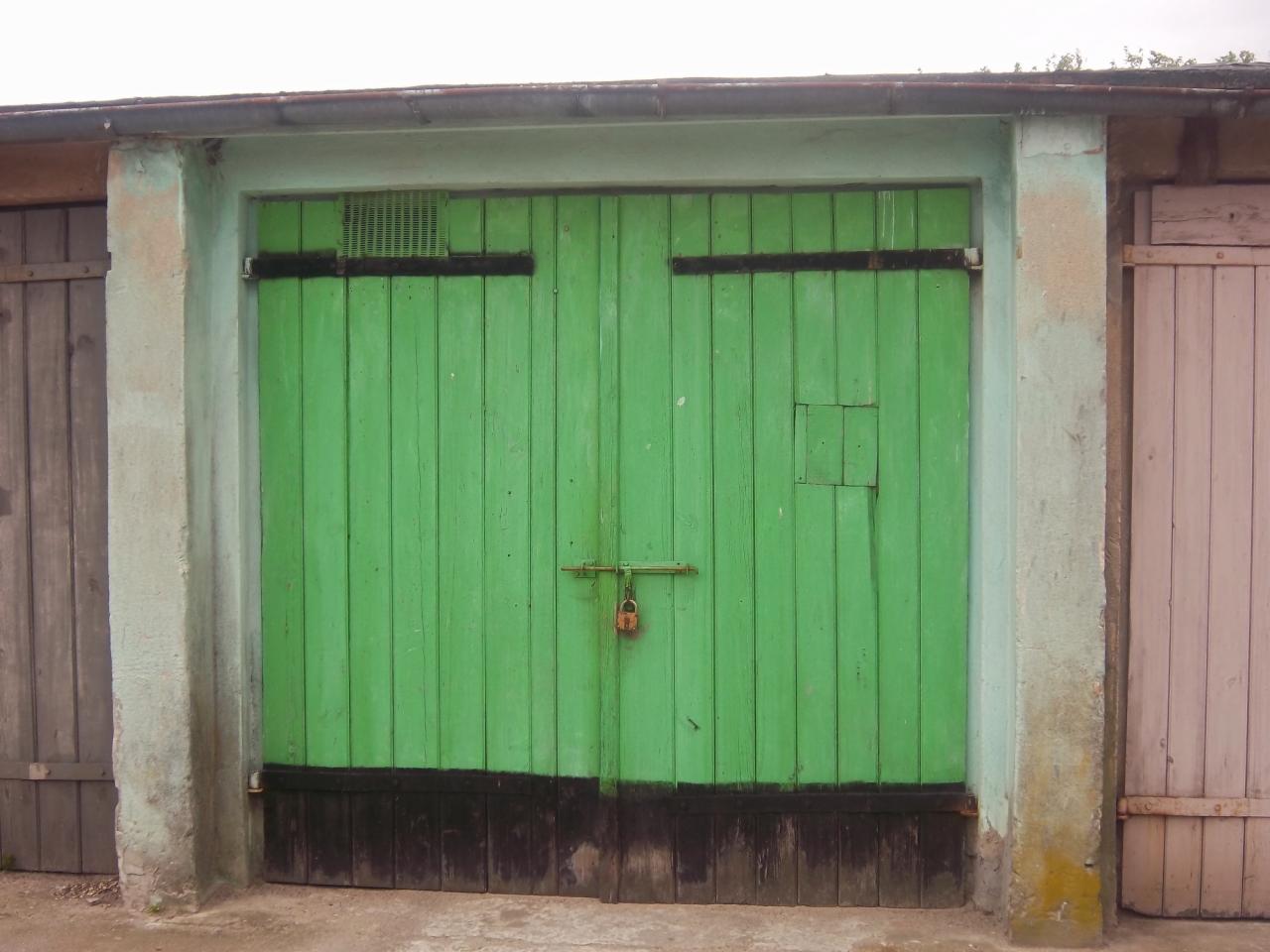 Zielone, dwuskrzydłowe drzwi garażowe z czarnymi akcentami i rdzawą kłódką, umieszczone pomiędzy sąsiednimi garażami o różnej kolorystyce.