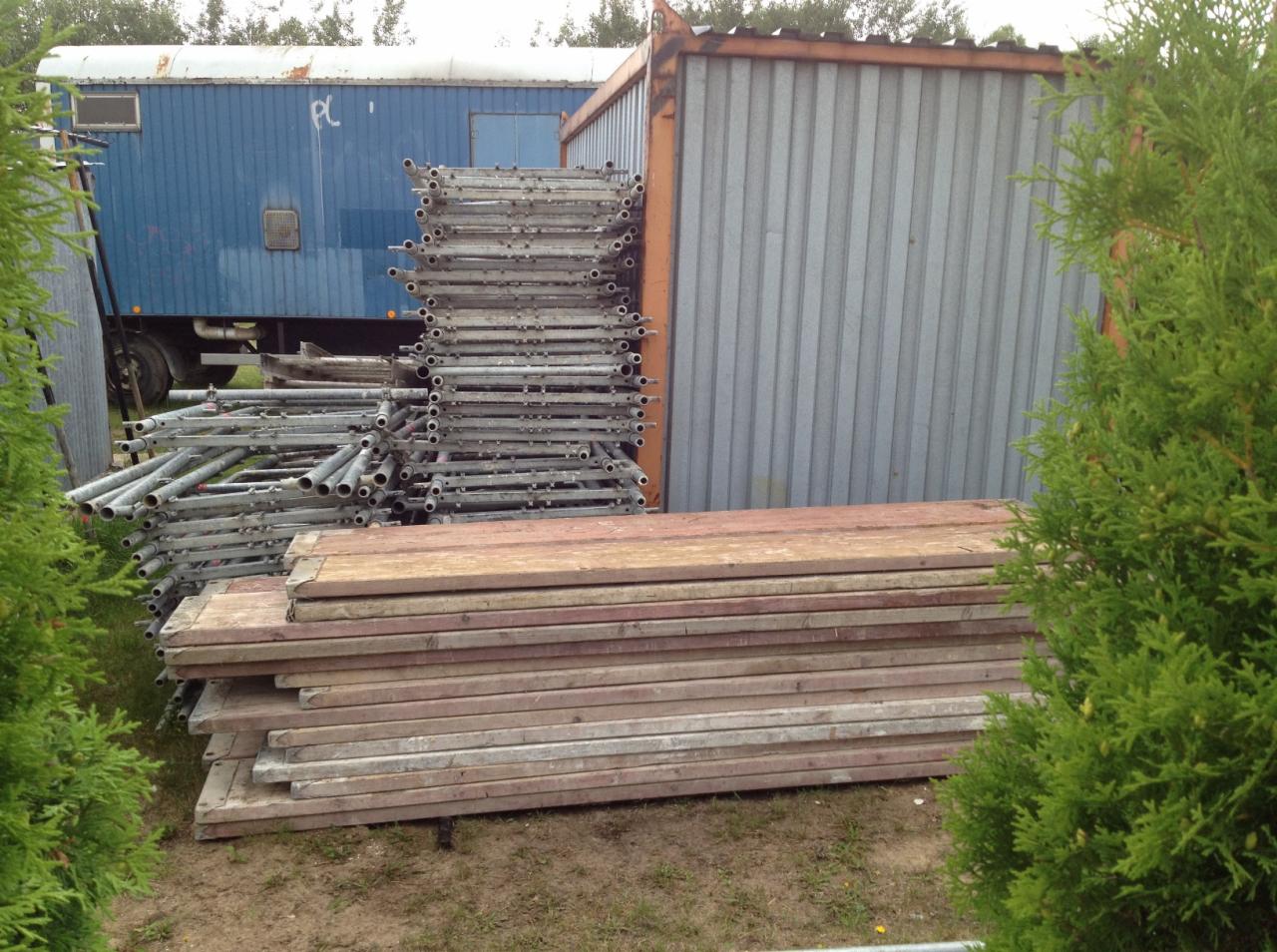 Stos rusztowania Plettac z metalowymi elementami i drewnianymi podestami, zaparkowany obok niebieskiego kontenera budowlanego i szarej blaszanej konstrukcji, z fragmentami zielonych krzewów po...