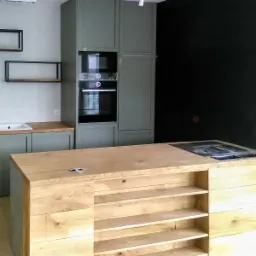 Nowoczesna kuchnia z wyspą z jasnego drewna i szarymi szafkami, czarna ściana w tle, widoczne półki i wbudowany piekarnik.