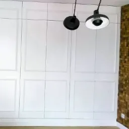 Biała szafa wnękowa z dekoracyjnymi panelami, oświetlona dwoma czarnymi lampami wiszącymi, uzupełniona ścianą z drewnianych paneli w Poznaniu.