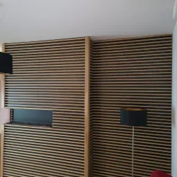 Drewniana ścianka działowa z poziomych listew w kolorze jasnego drewna i ciemnego brązu, oddzielająca przestrzeń, z widocznymi lampami po obu stronach.