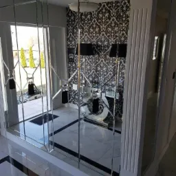 Nowoczesna ściana z lustrzanych paneli z geometrycznym wzorem, białe ramy, widok na przedpokój z marmurową podłogą i wzorzystą tapetą.