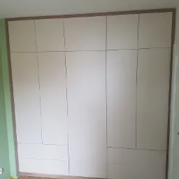 Wbudowana szafa z jasnymi frontami i drewnianym obramowaniem, dopasowana do ściany, widoczna na tle parkietu i zielonej ściany.