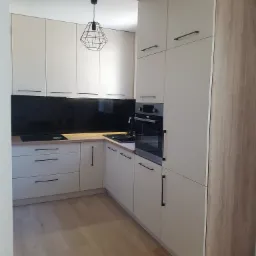 Nowoczesna kuchnia z beżowymi szafkami z czarnymi uchwytami, czarnym panelem szklanym nad blatem i drewnianą podłogą, oświetlona geometryczną lampą wiszącą.