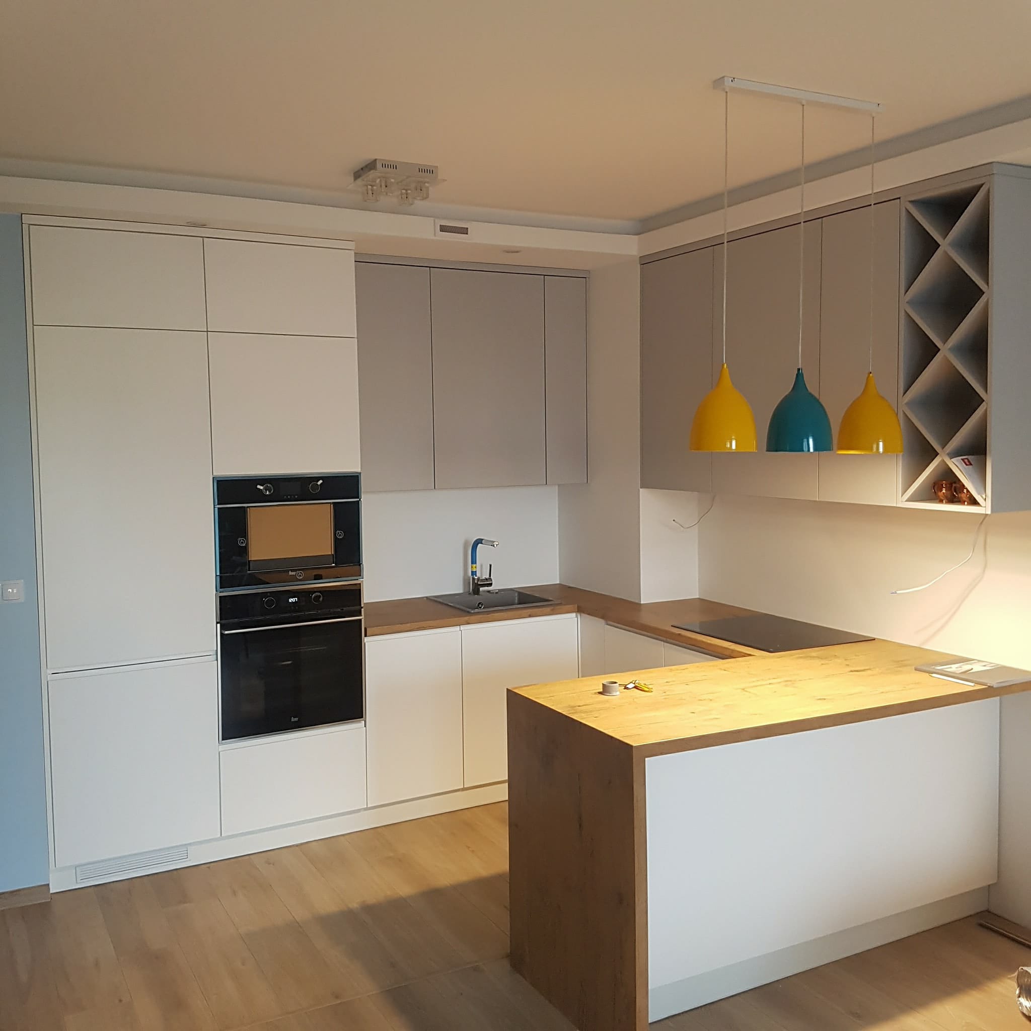 Nowoczesna kuchnia z białymi i szarymi szafkami, drewnianym blatem i trzema kolorowymi lampami wiszącymi: żółtą i turkusową.