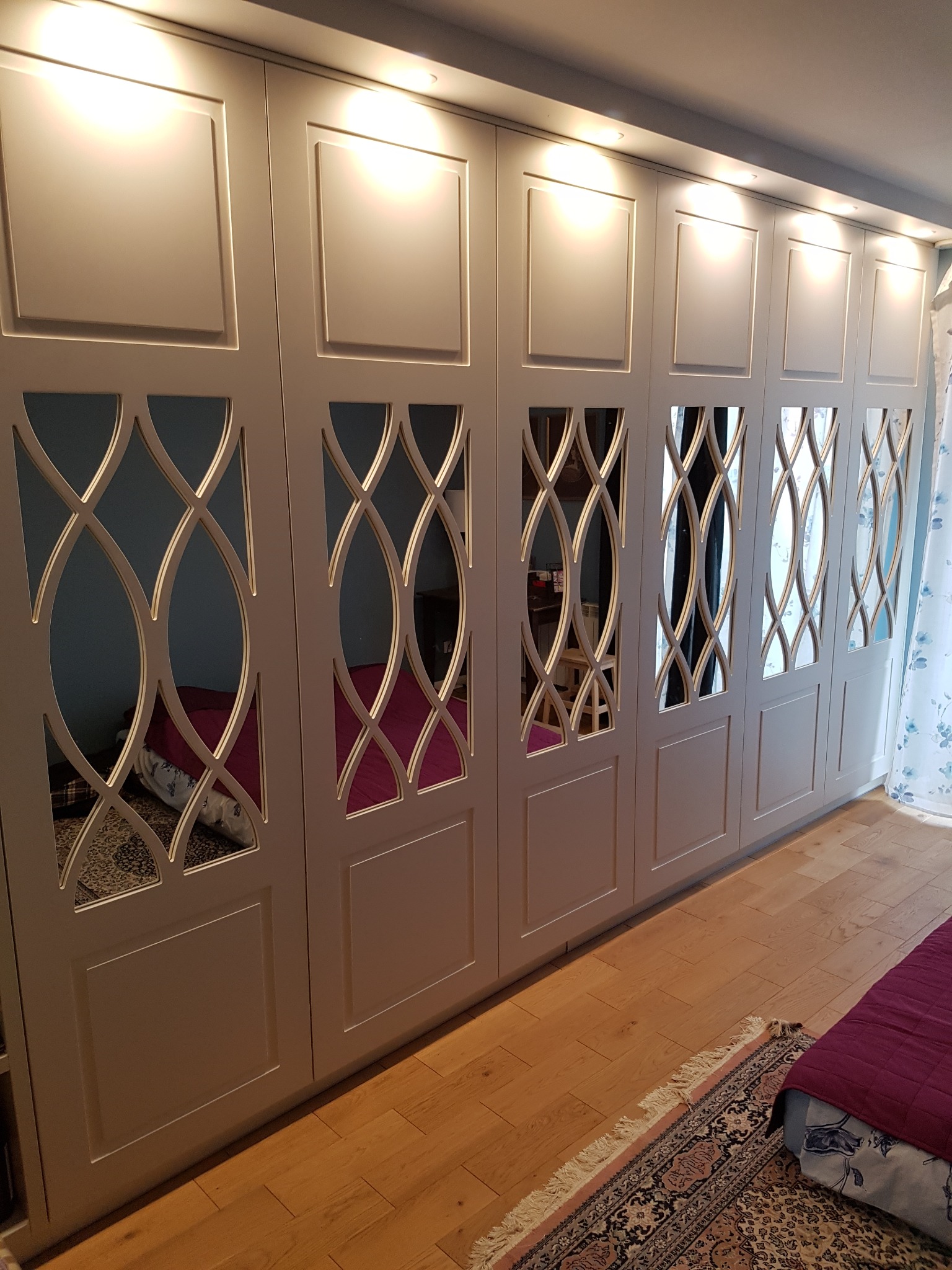 Długa, jasnoszara szafa z dekoracyjnymi, ażurowymi wstawkami, oświetlona od góry punktowymi lampami, widoczna w sypialni z parkietem i fragmentem łóżka z bordową narzutą