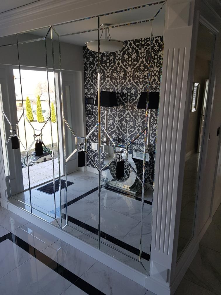 Nowoczesna ściana z lustrzanych paneli z geometrycznym wzorem, białe ramy, widok na przedpokój z marmurową podłogą i wzorzystą tapetą.