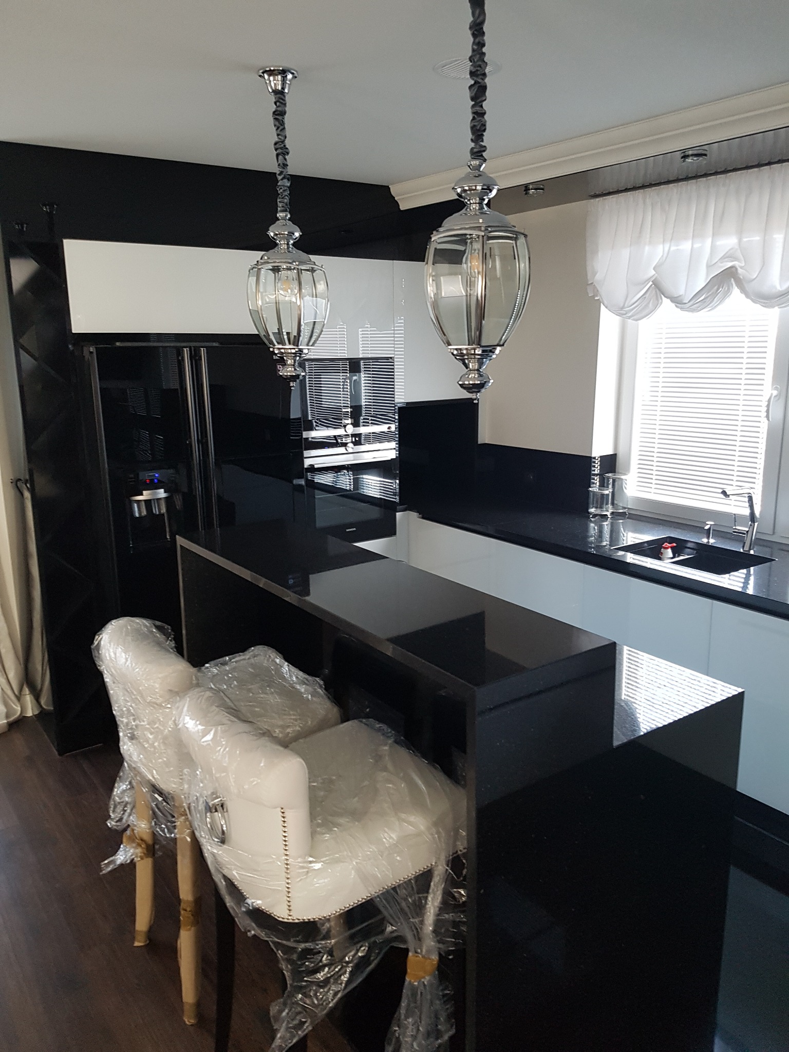 Nowoczesna kuchnia z czarno-białymi szafkami, blatem z ciemnego granitu i dwoma wiszącymi lampami w stylu retro; krzesła barowe w folii ochronnej.