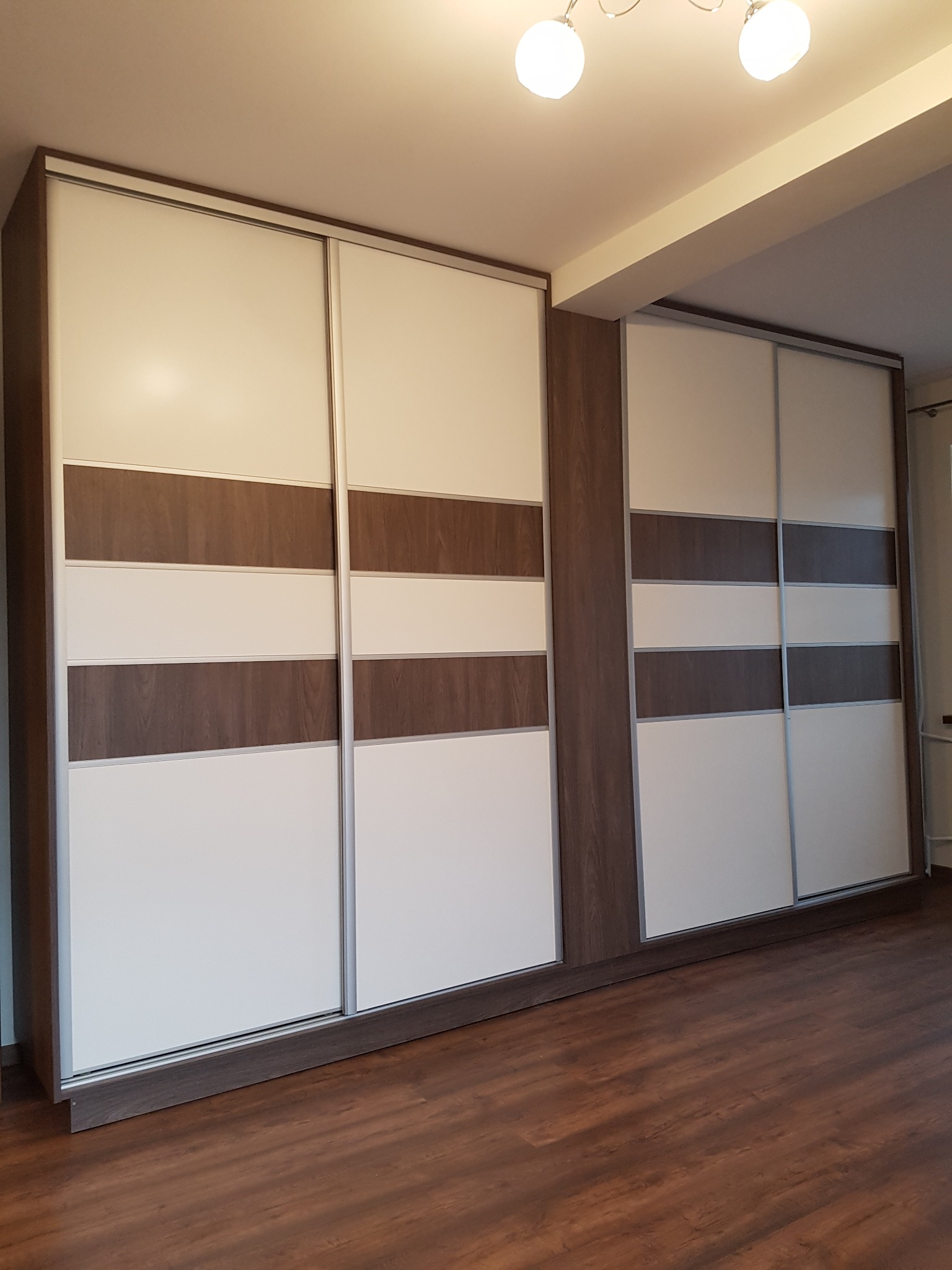 Dwie duże szafy z przesuwnymi drzwiami w kolorze białym i drewnopodobnym, z widocznym podziałem na poziome panele, na tle drewnianej podłogi i białego sufitu z lampami.