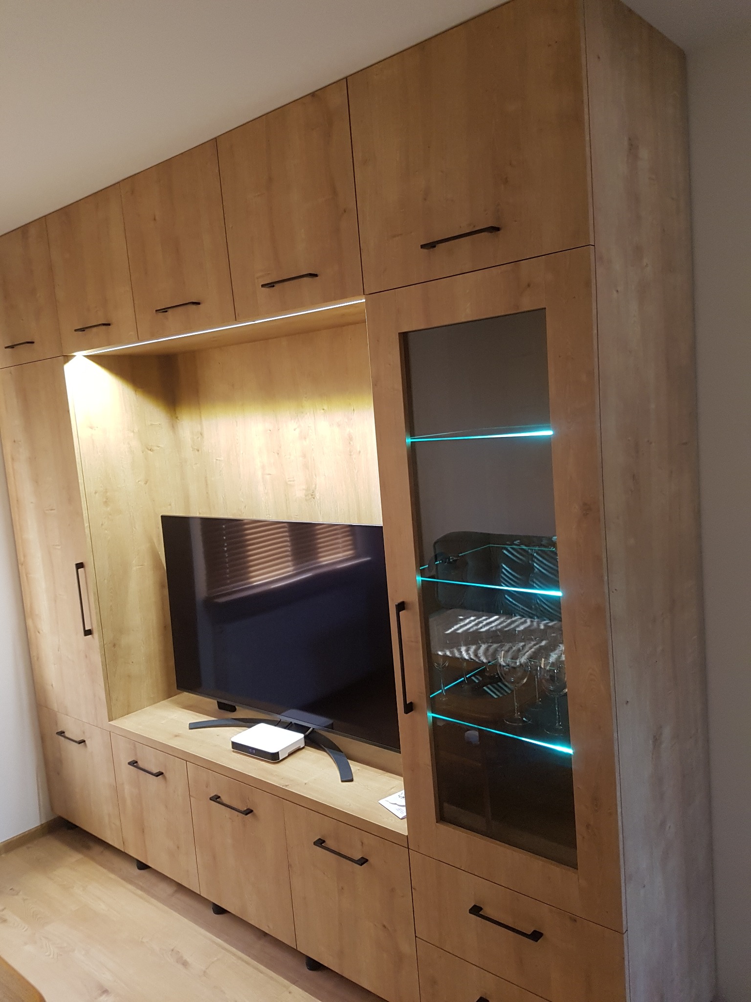 Zabudowa meblowa w salonie z frontami w odcieniach jasnego drewna, czarne, podłużne uchwyty, przeszklona witryna z podświetleniem LED, telewizor w centralnej części.