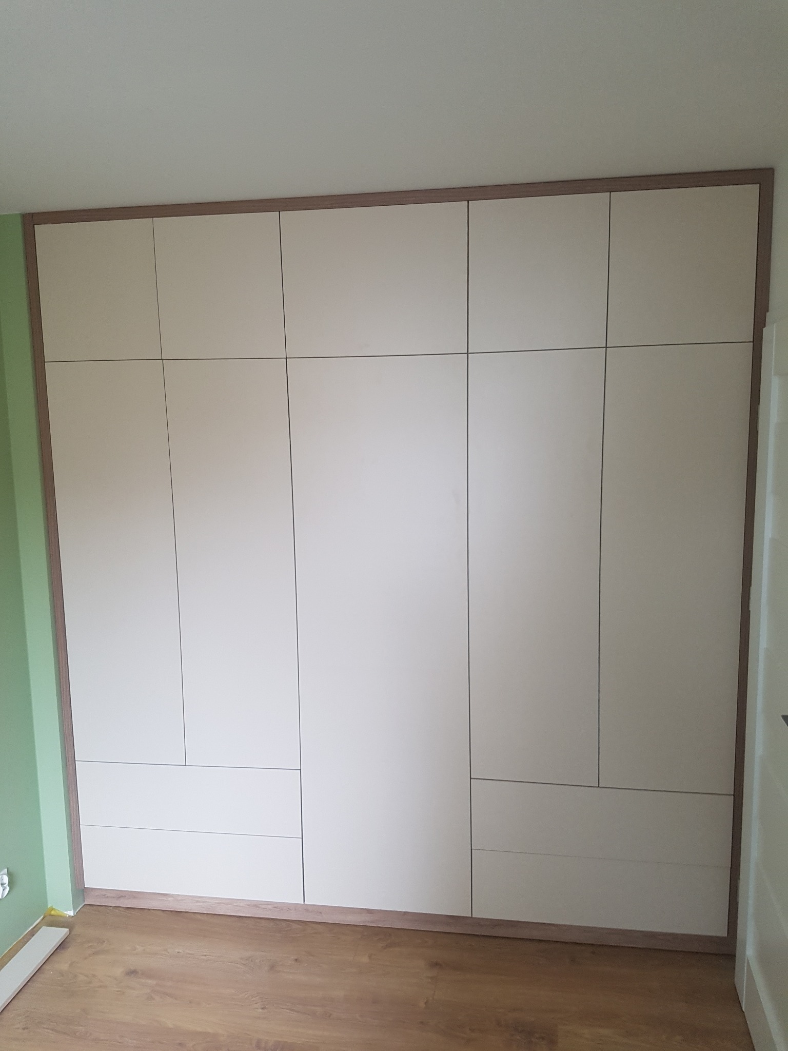 Wbudowana szafa z jasnymi frontami i drewnianym obramowaniem, dopasowana do ściany, widoczna na tle parkietu i zielonej ściany.