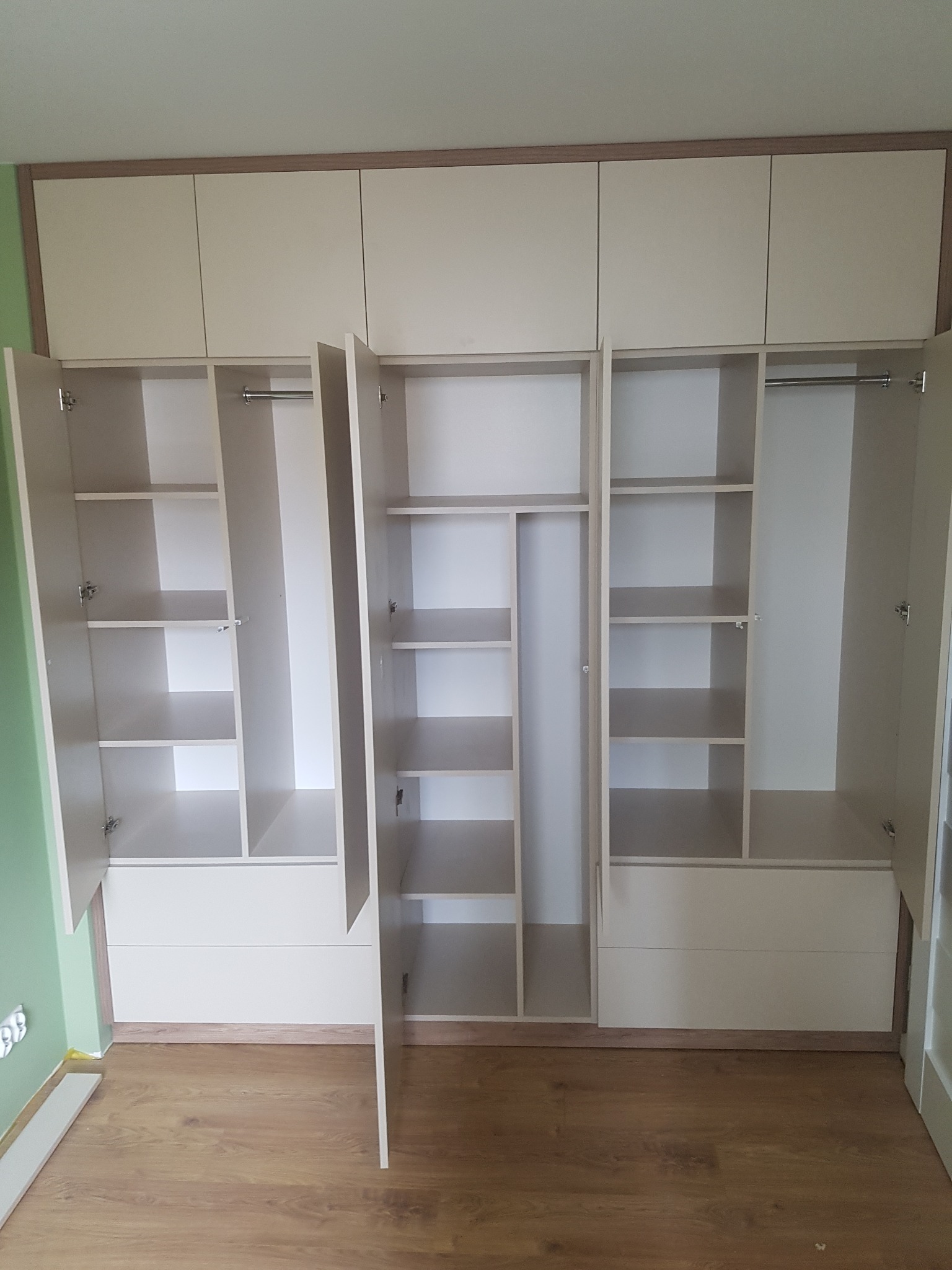 Jasna zabudowa meblowa z otwartymi drzwiami, ukazująca półki i szuflady, z widoczną podłogą z paneli i fragmentem zielonej ściany.