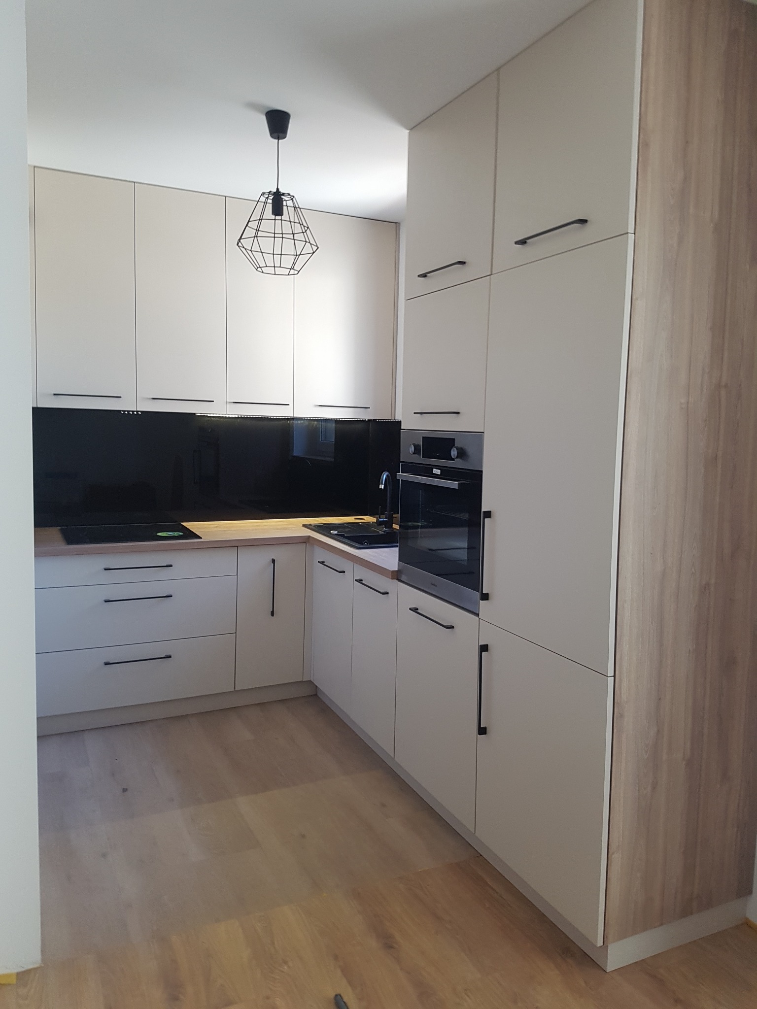 Nowoczesna kuchnia z beżowymi szafkami z czarnymi uchwytami, czarnym panelem szklanym nad blatem i drewnianą podłogą, oświetlona geometryczną lampą wiszącą.