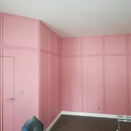 Ściana w pokoju pomalowana na różowo z dekoracyjnymi panelami ściennymi i drzwiami w tym samym kolorze, widoczny fragment podłogi w jodełkę i fragment drabiny malarskiej.