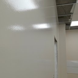 Gipsiorz-Malyrz.itp - Świeżo pomalowana, błyszcząca ściana w biurze, widoczny fragment sufitu podwieszanego i ościeżnica drzwi w trakcie wykończenia.