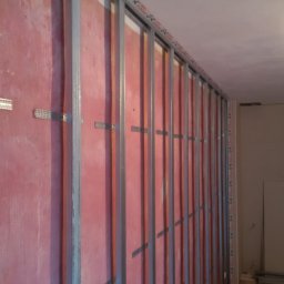 Gipsiorz-Malyrz.itp - Metalowy szkielet konstrukcji ścianki działowej z profili GK na tle czerwonej ściany, widoczne elementy mocujące i oznaczenia producenta profili.