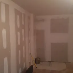 Wnętrze w trakcie prac wykończeniowych: ściany z nałożonymi pasami gładzi, widoczny wałek malarski oparty o ścianę, wiadro z masą szpachlową i fragment skrzynki narzędziowej na podłodze.