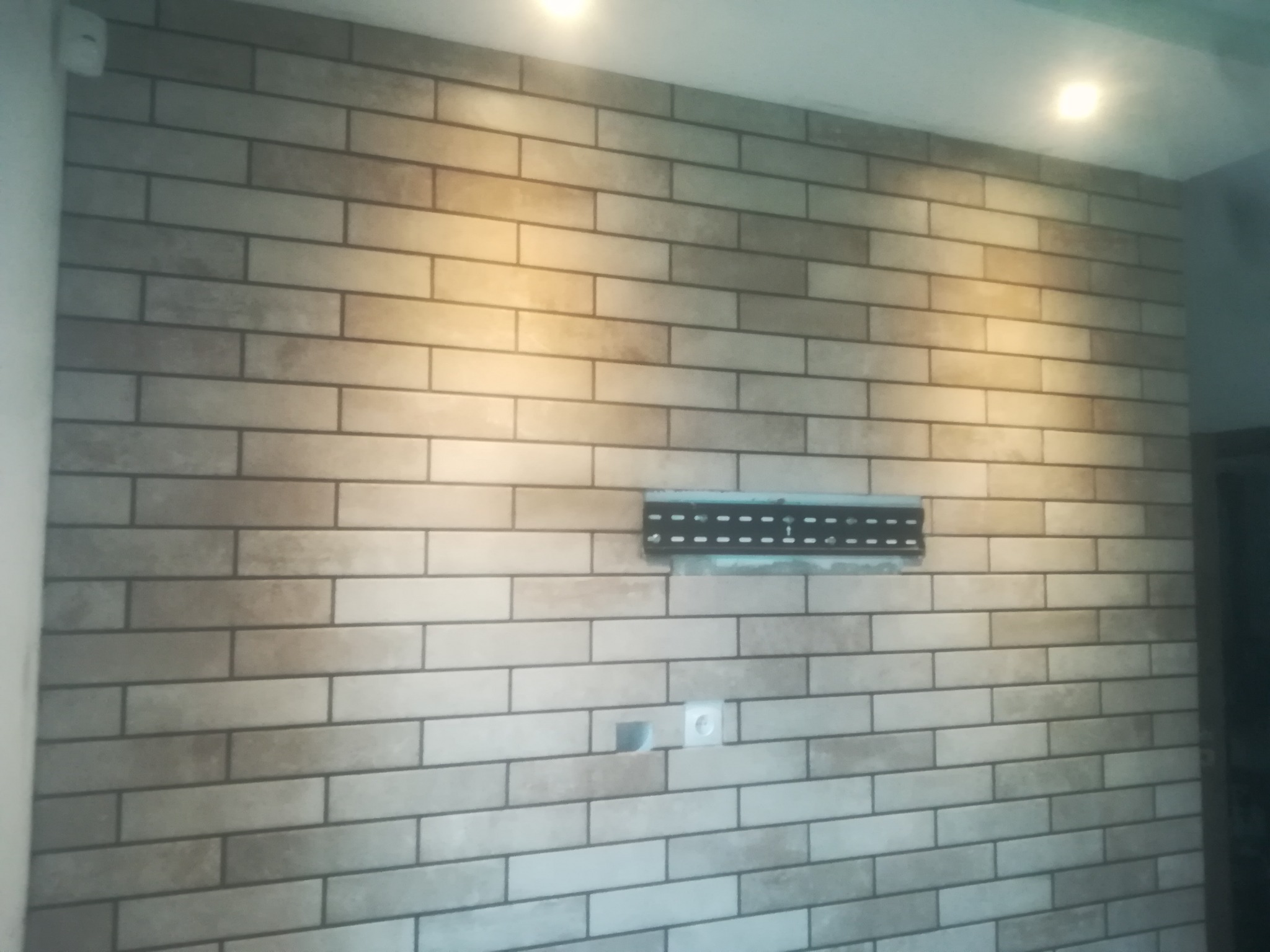 Ściana wyłożona płytkami imitującymi cegłę w odcieniach beżu i szarości, z zamontowanym uchwytem do telewizora i gniazdkiem elektrycznym.