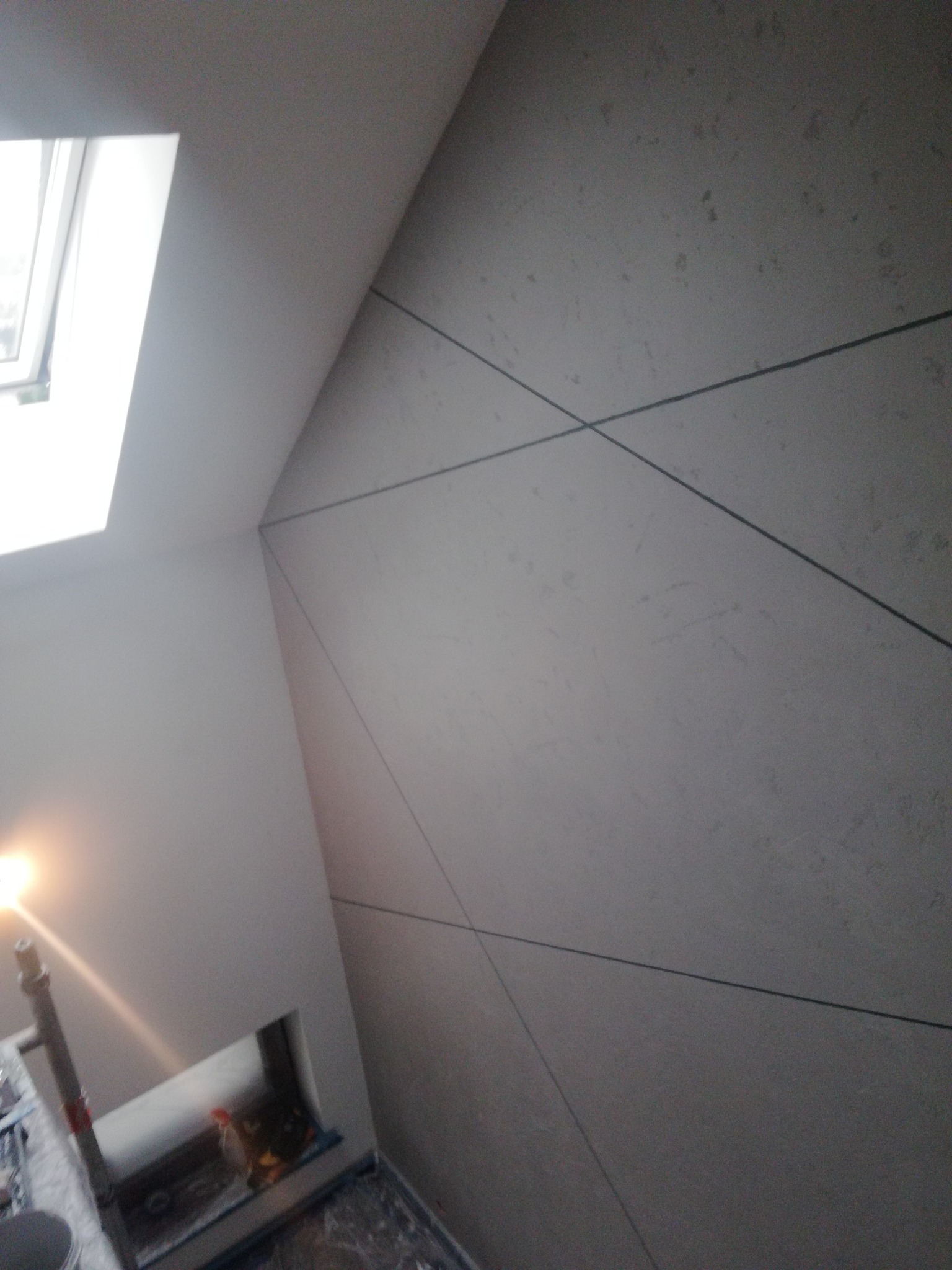 Skośna ściana poddasza z oknem dachowym, wykończona jasną gładzią z ciemnymi liniami tworzącymi geometryczny wzór. Widoczne narzędzia malarskie i zabezpieczona folią podłoga.