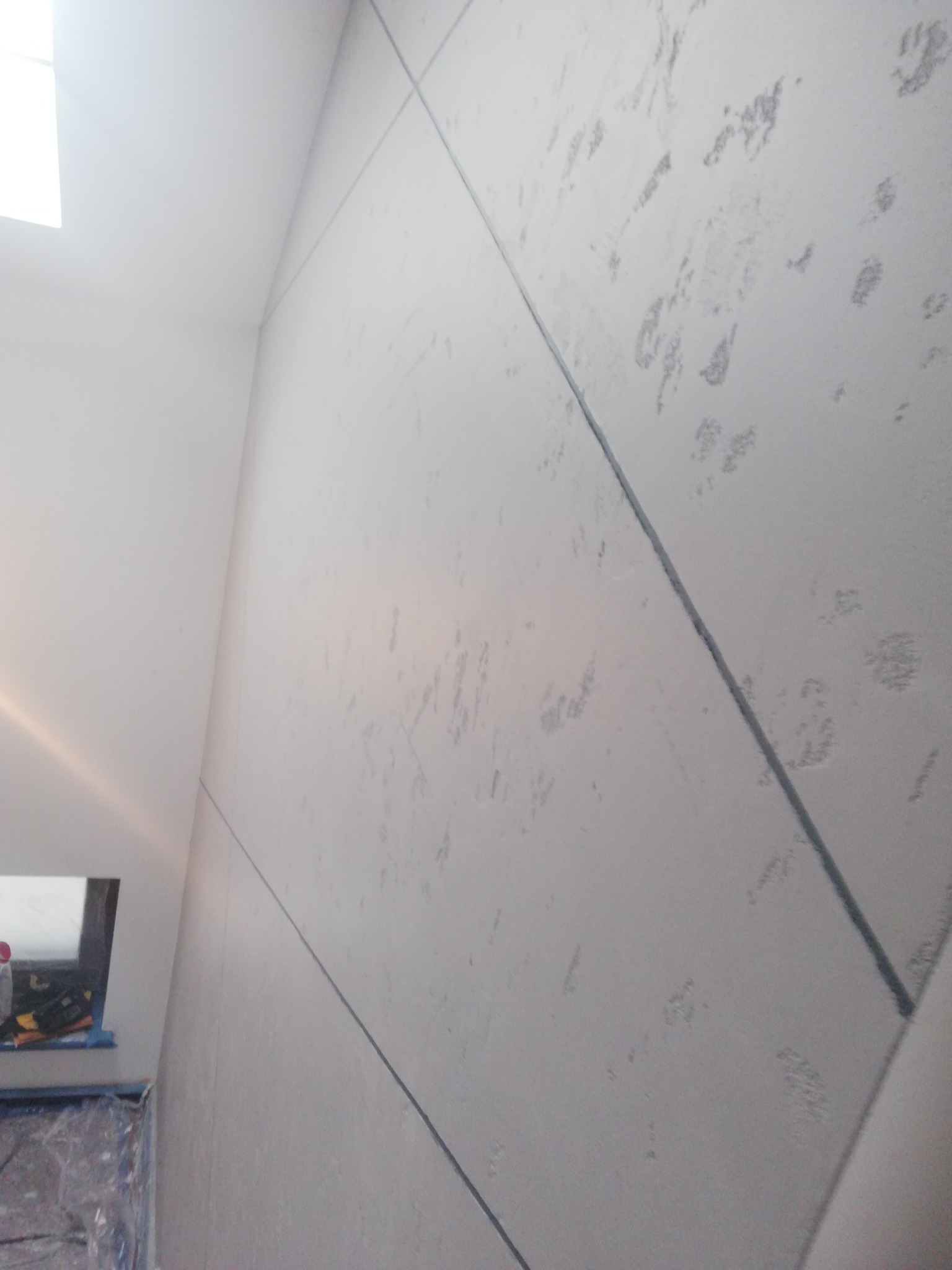 Ściana pokryta dekoracyjną, szarą strukturą z wyraźnymi podziałami na panele, widoczny fragment okna w lewym górnym rogu i zabezpieczone folią malarską otoczenie.