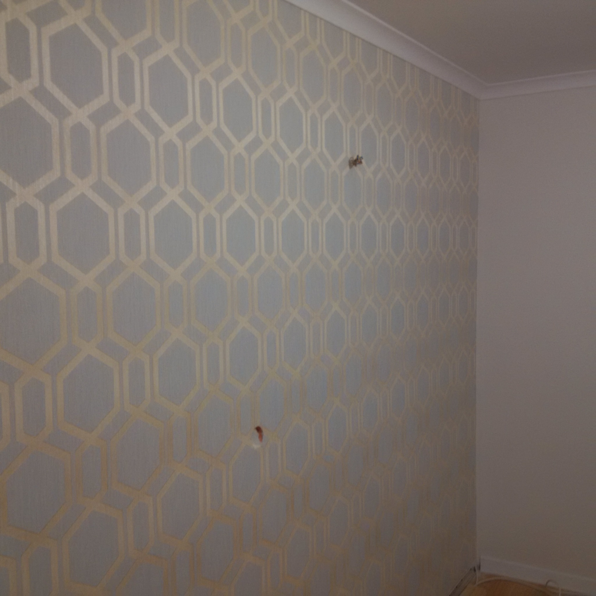 Ściana z szarą tapetą w złote geometryczne wzory; widoczne dwa otwory po kołkach, listwa przypodłogowa i sufitowa.