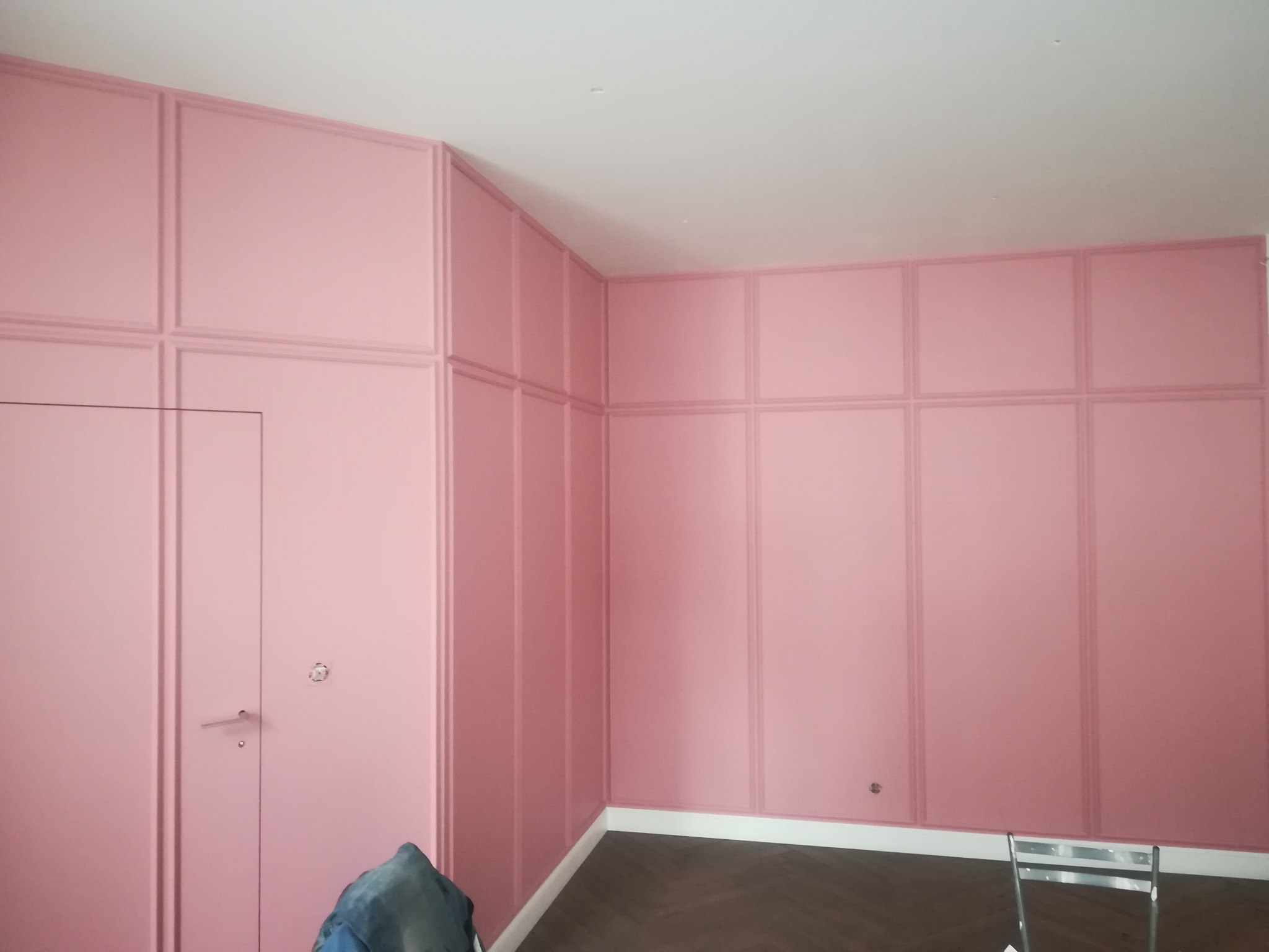 Ściana w pokoju pomalowana na różowo z dekoracyjnymi panelami ściennymi i drzwiami w tym samym kolorze, widoczny fragment podłogi w jodełkę i fragment drabiny malarskiej.