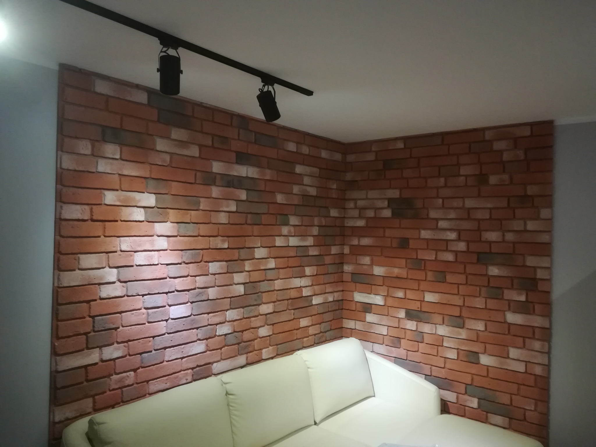 Ściana wykończona cegłą dekoracyjną w salonie z kremową sofą i oświetleniem punktowym na suficie.