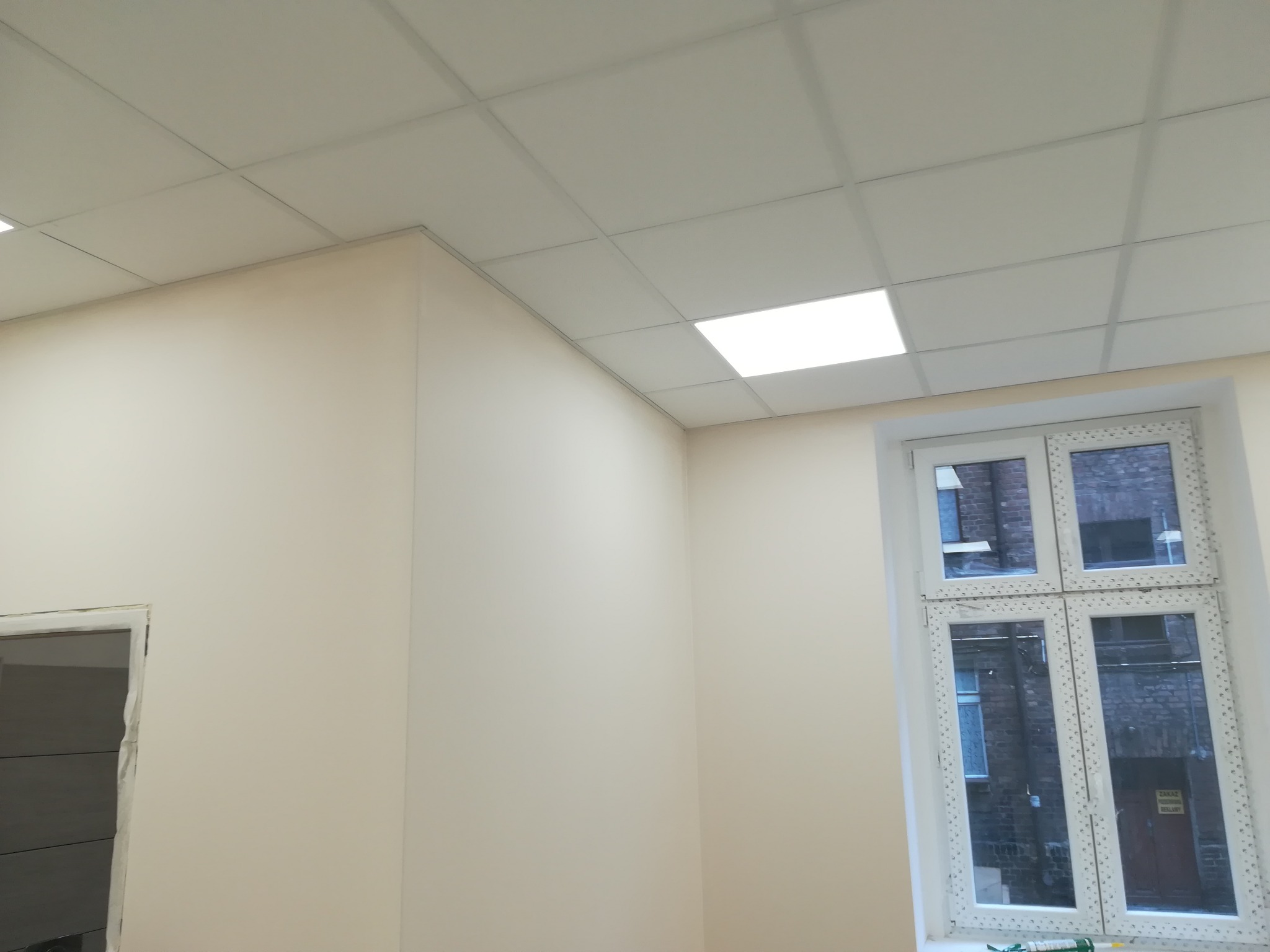 Wnętrze biura po remoncie: kremowe ściany, sufit podwieszany z kwadratowymi panelami, białe okno z widokiem na ceglany budynek. Widoczny częściowo otwór drzwiowy.