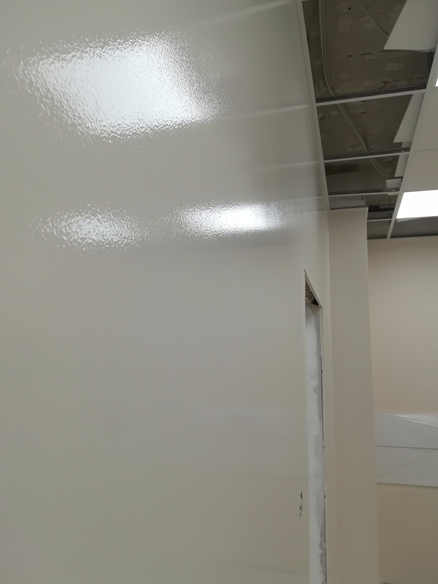 Świeżo pomalowana, błyszcząca ściana w biurze, widoczny fragment sufitu podwieszanego i ościeżnica drzwi w trakcie wykończenia.