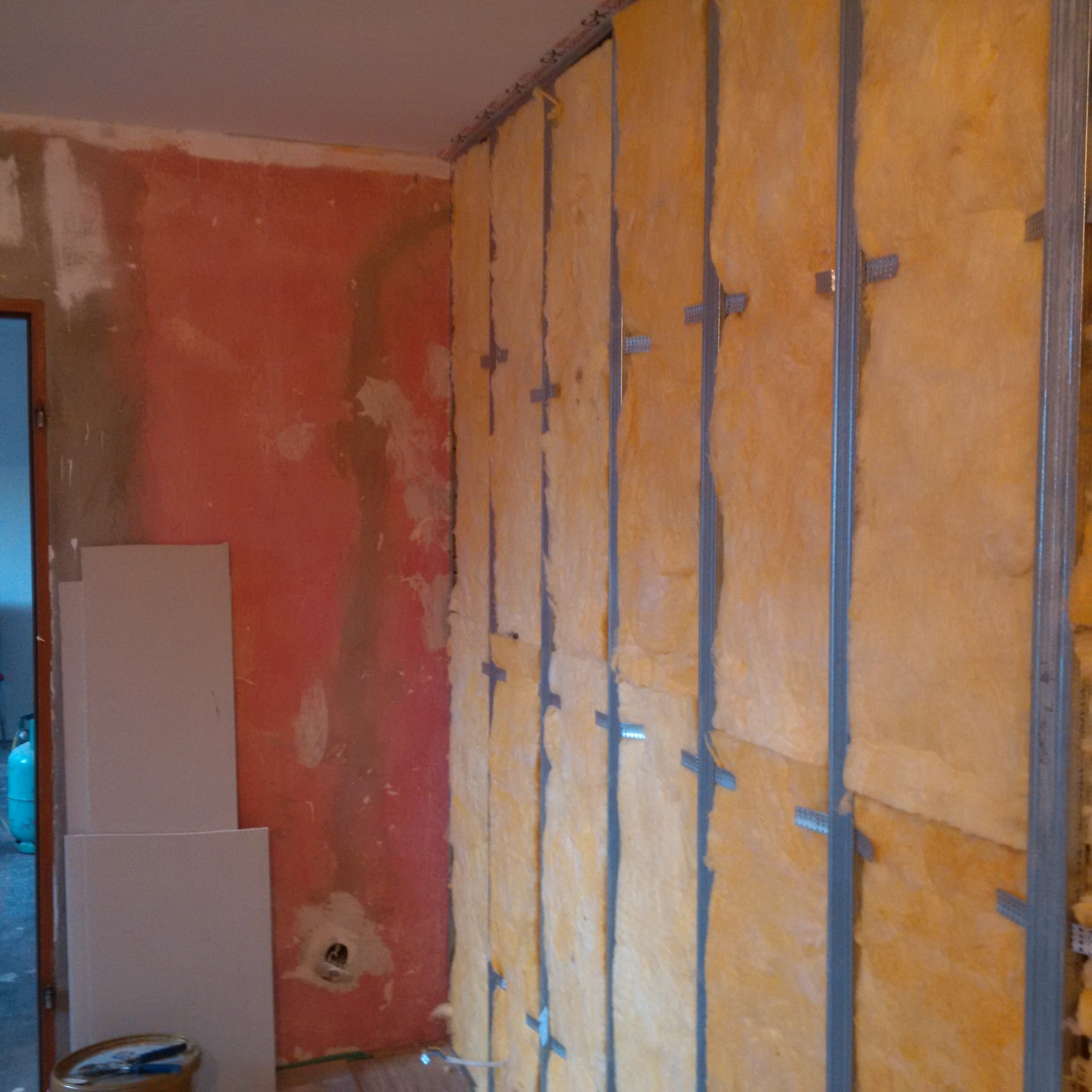 Montaż izolacji z wełny mineralnej na ścianie z metalowym stelażem, obok fragment ściany z resztkami starej farby i przygotowane płyty kartonowo-gipsowe.