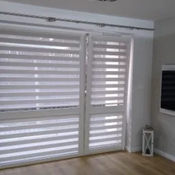 Dwuskrzydłowe okno balkonowe z zamontowanymi roletami dzień-noc w białych ramach, widoczne na tle jasnego wnętrza z drewnianą podłogą i ścianą z telewizorem.