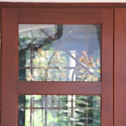 Dwuskrzydłowe drzwi wewnętrzne z brązowego drewna z szybami z geometrycznymi szprosami, widoczne zawiasy i odbicie zieleni z zewnątrz.