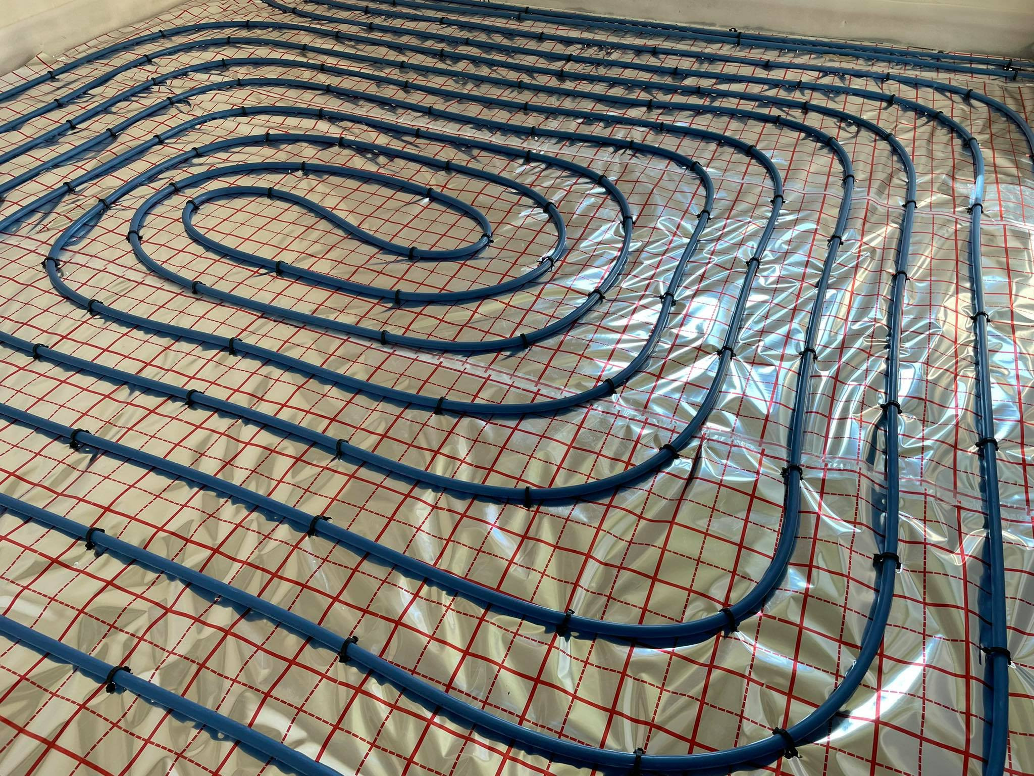 Niebieskie rury ogrzewania podłogowego ułożone spiralnie na srebrnej folii z czerwoną kratką, zabezpieczone czarnymi opaskami zaciskowymi.