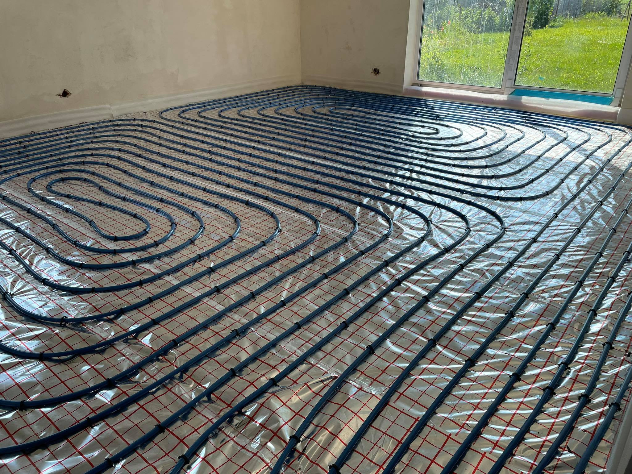 Rozłożone, niebieskie rury ogrzewania podłogowego na folii z czerwoną kratką w surowym pomieszczeniu z oknem i widokiem na zieleń.