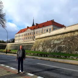 Mężczyzna w skórzanej kurtce stoi na chodniku przed barokowym pałacem z czerwonym dachem i kamiennym murem obronnym.