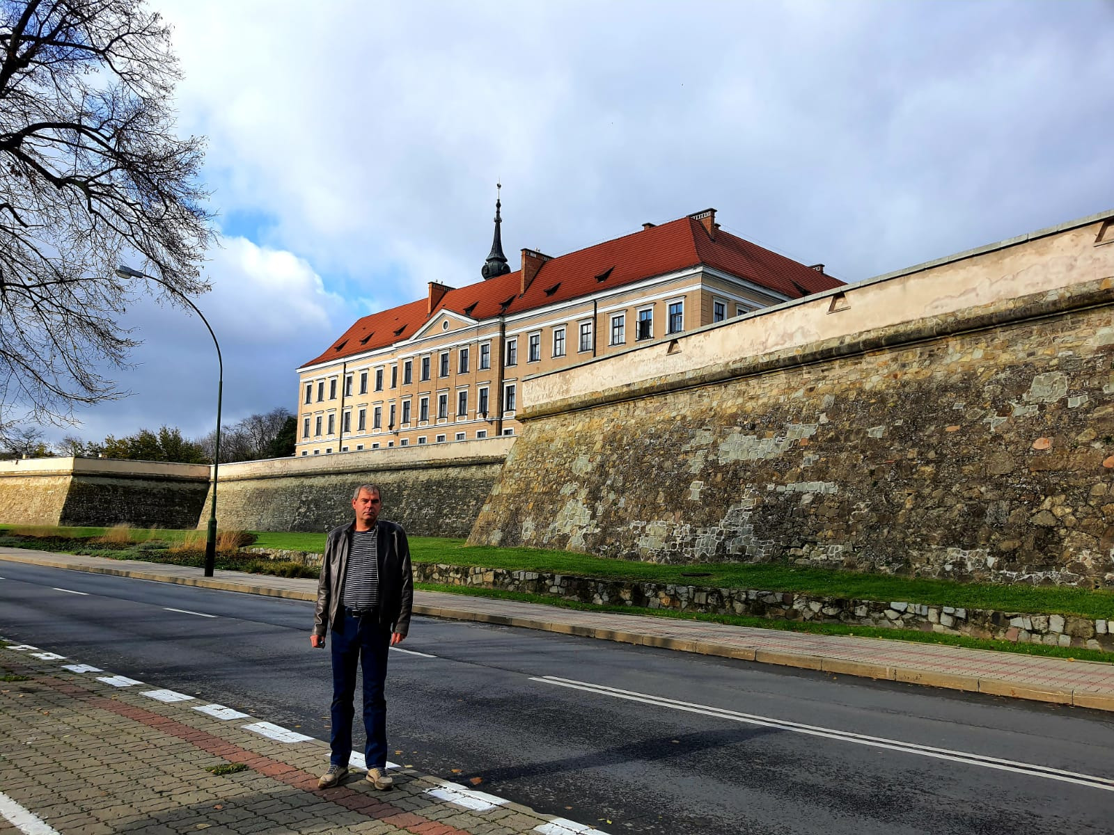 Mężczyzna w skórzanej kurtce stoi na chodniku przed barokowym pałacem z czerwonym dachem i kamiennym murem obronnym.