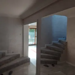 Klatka schodowa z wykończonymi ścianami w odcieniach beżu i szarości, obłożona wykładziną dywanową, widoczna z perspektywy dolnej części schodów, z otwartym przejściem do jasnego pomieszczenia w tle.